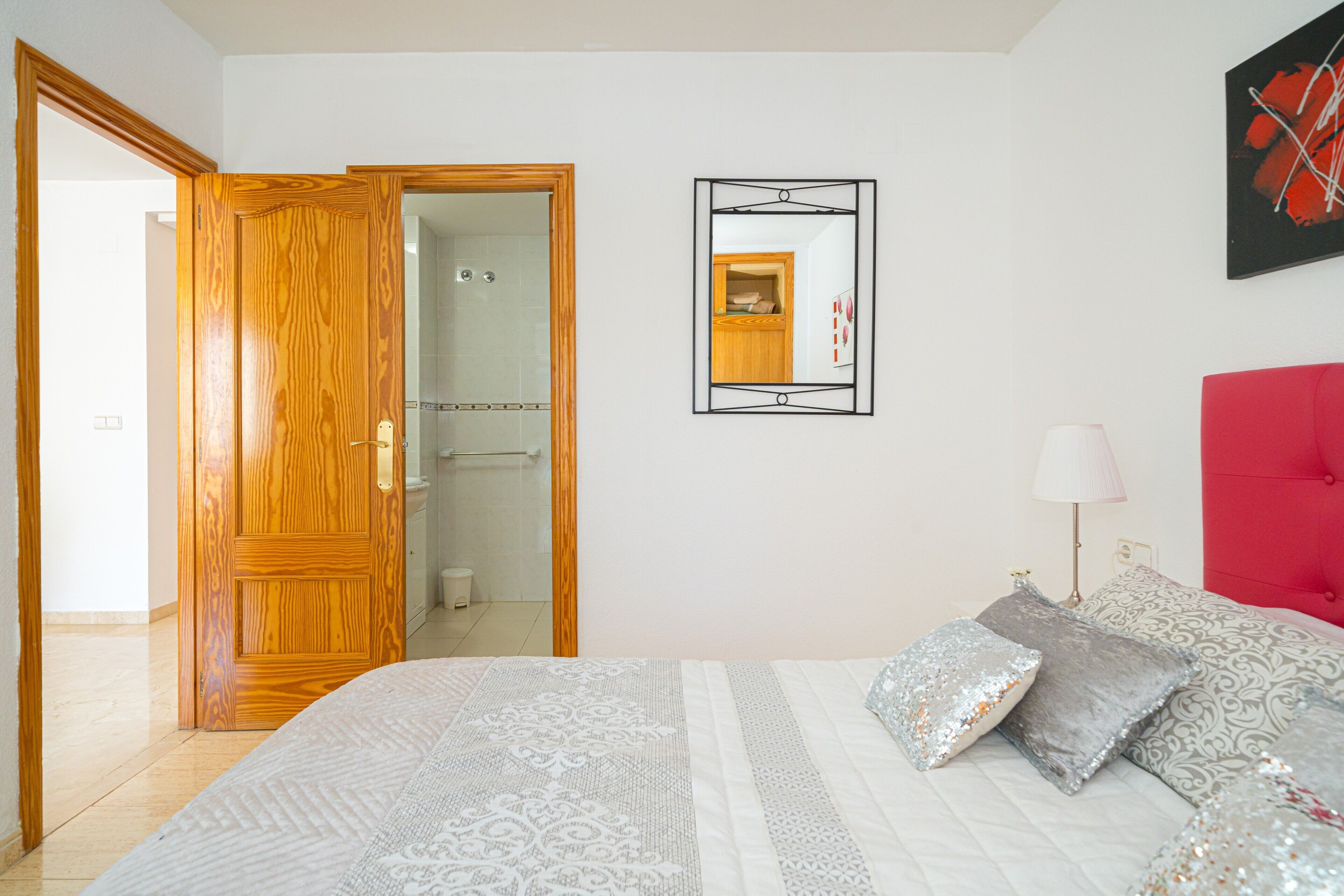 Ein Schlafzimmer mit einem Bett, Kissen, einer Nachttischlampe, einem Spiegel und einer Tür zu einem Badezimmer.