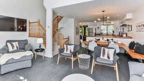 A modern living room with grey sofas, a wooden staircase, and a kitchen area with a dining table. - 4 Storey Ski In/Out Mountain Home in Tremblant (Mont-Tremblant)