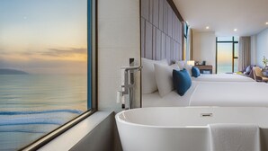 A modern hotel room with a large window offering a view of the sea, a large bathtub, and a neatly made bed with pillows.