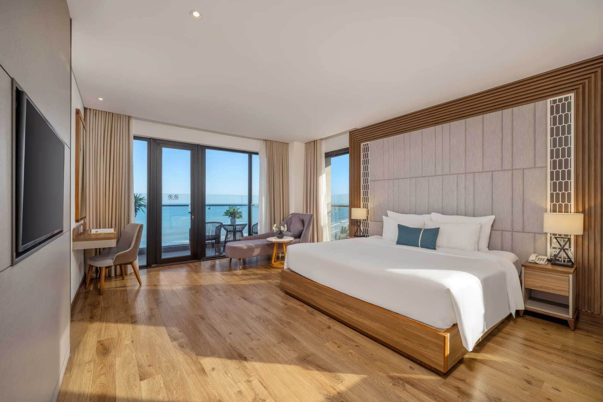 A spacious hotel room with a large bed, a desk, and a balcony with a view of the sea.