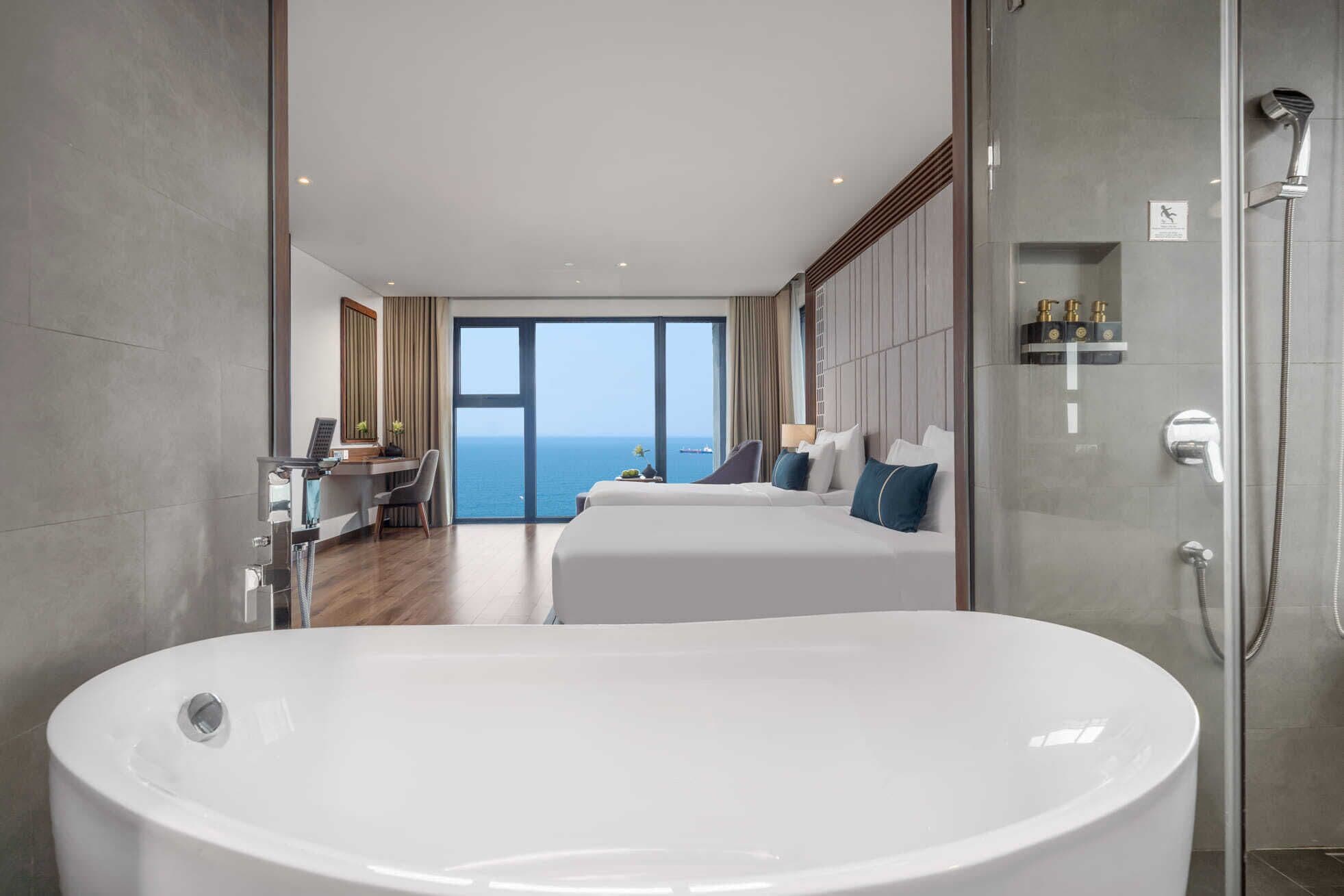 A modern hotel room with a large bathtub, a desk with a computer, and a view of the sea.