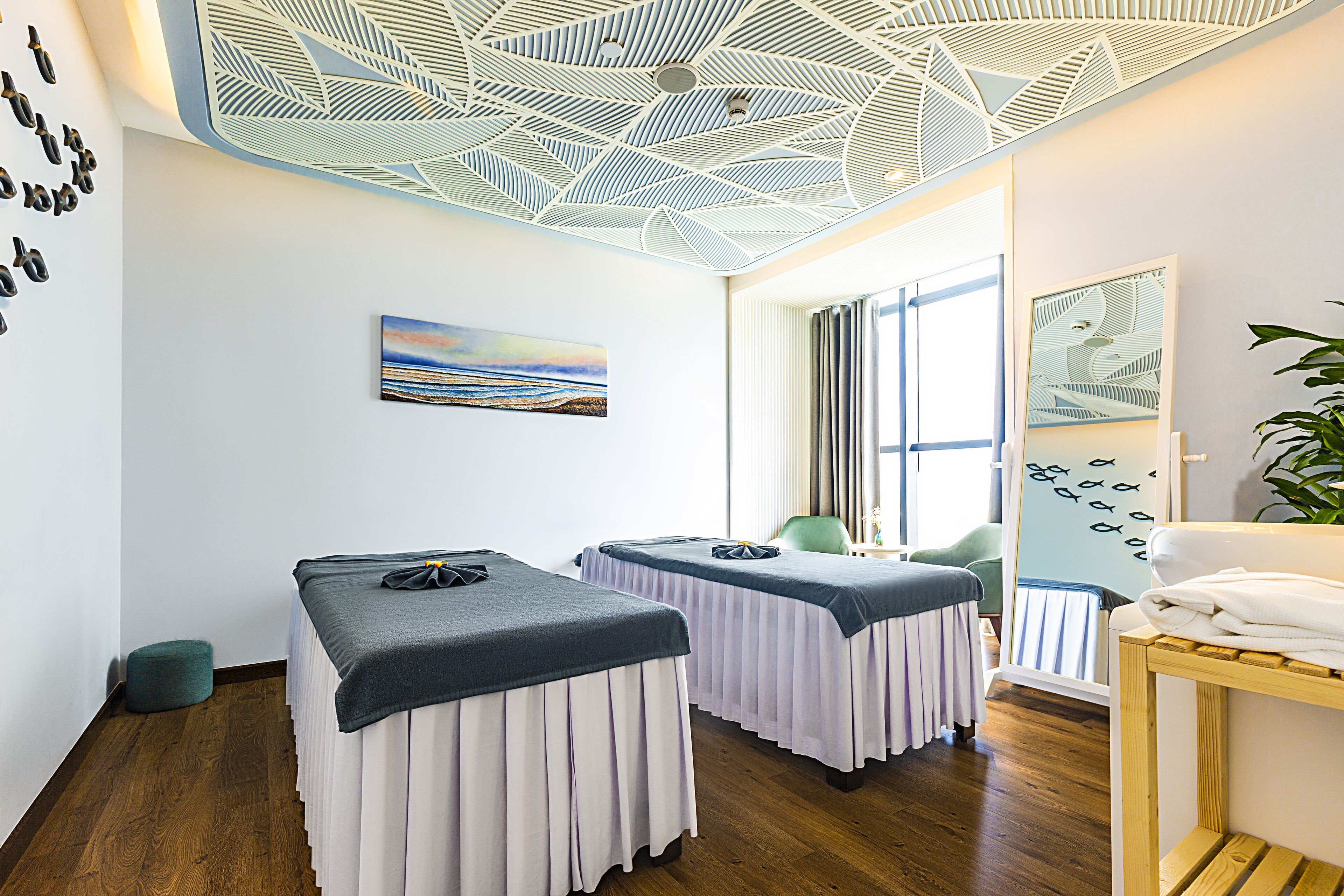A room with two massage tables, a wooden floor, a ceiling with a geometric pattern, a painting of a landscape, and a window with curtains.