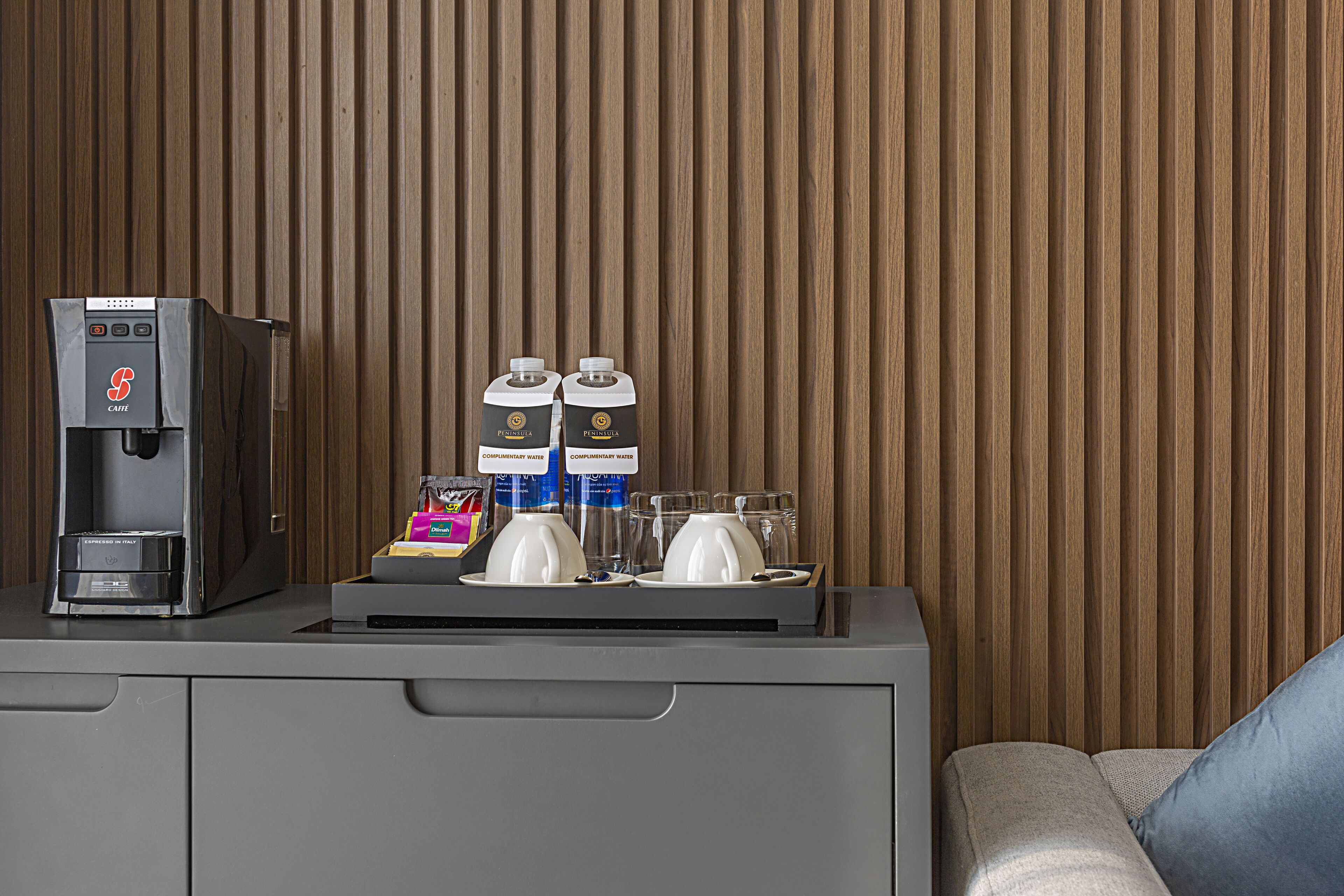 A coffee machine, two cartons of complimentary water, and a tray with two cups and two glasses on a counter against a wooden wall.