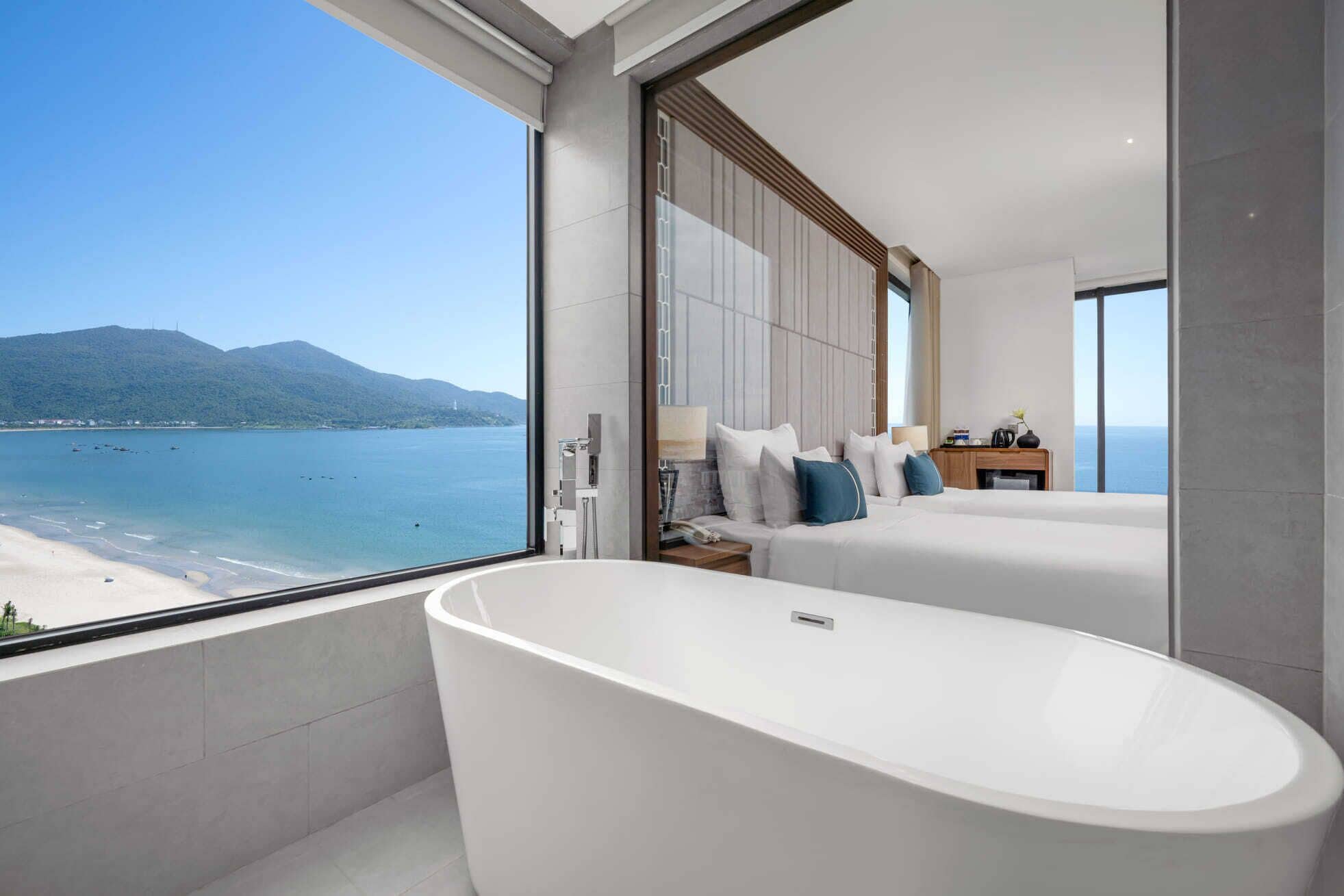Een moderne badkamer met een groot bad, een bed met wit beddengoed en uitzicht op zee en bergen.