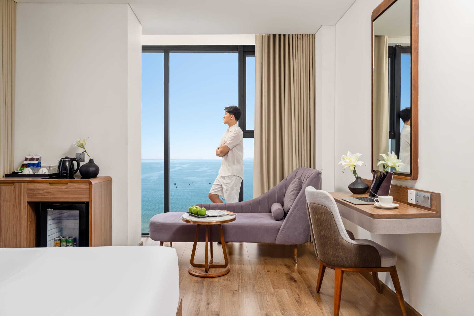 A hotel room with a bed, a desk with a laptop, a chair, a small table with a vase, and a view of the sea through a large window.