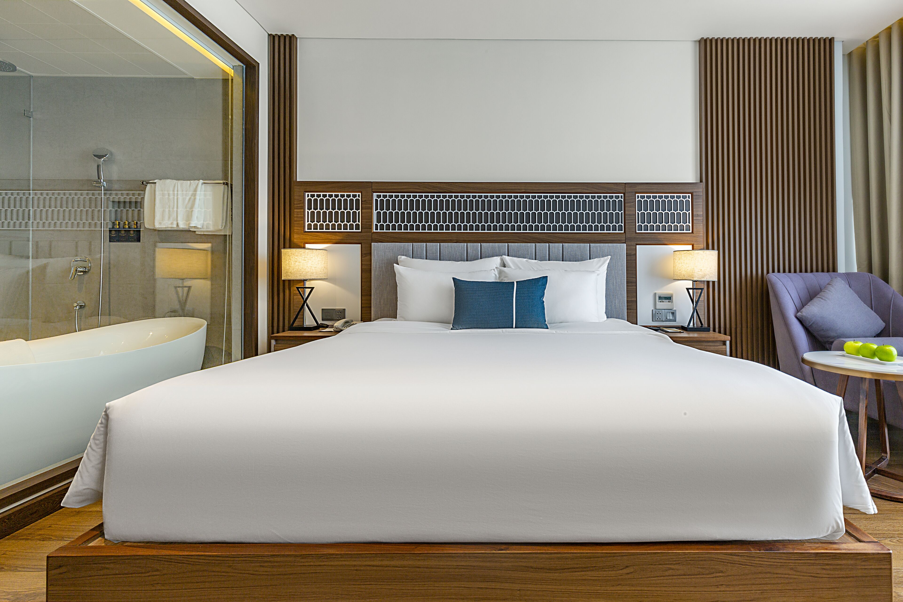A hotel room with a large bed, a bathtub, and a chair with apples on a side table.