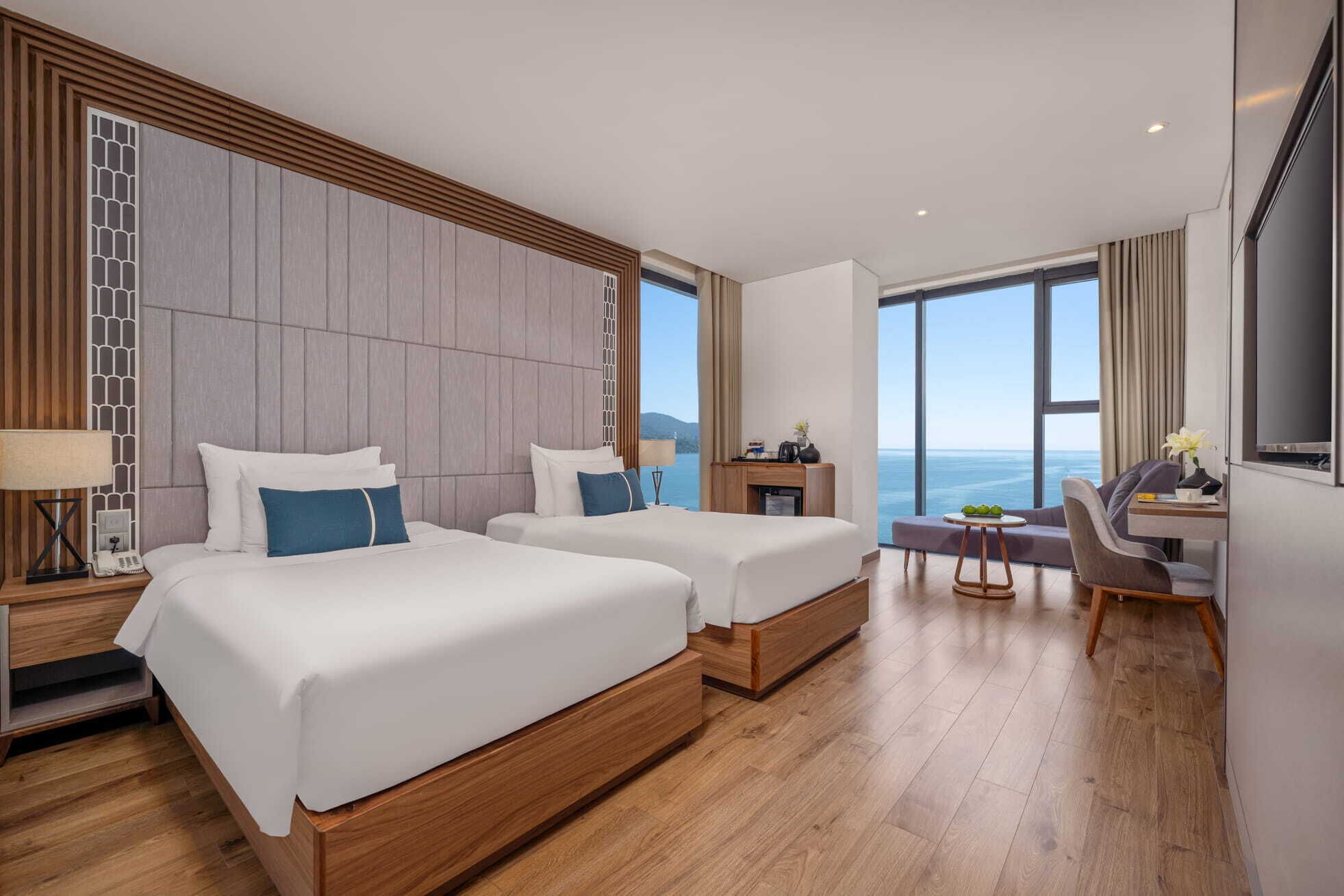 A hotel room with two beds, a desk, and a large window overlooking the sea.