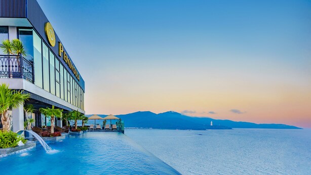 A hotel with a pool extending to the water, a mountain in the background, and a clear sky.