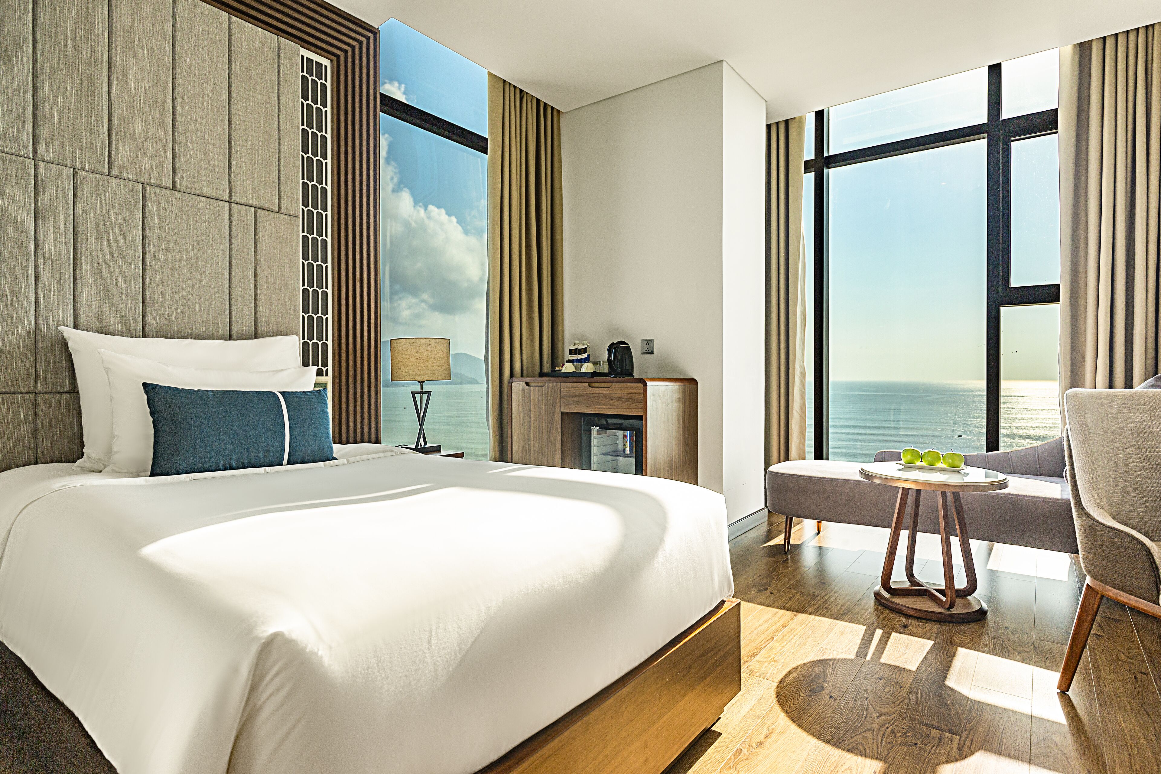 A modern hotel room with a large bed, a seating area with a table, and a view of the sea.