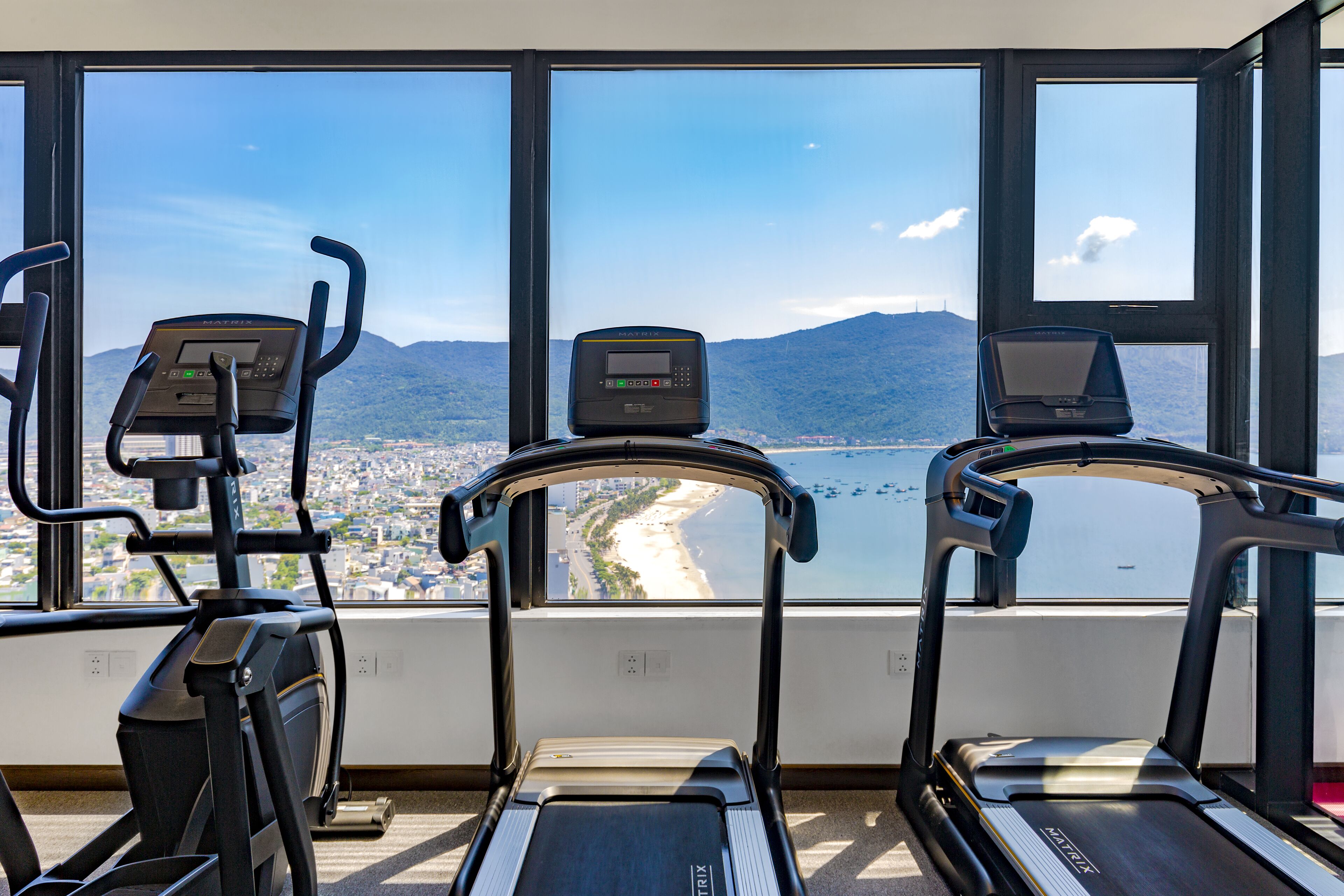 A gym with treadmills and exercise bikes, large windows with a view of a beach and mountains.