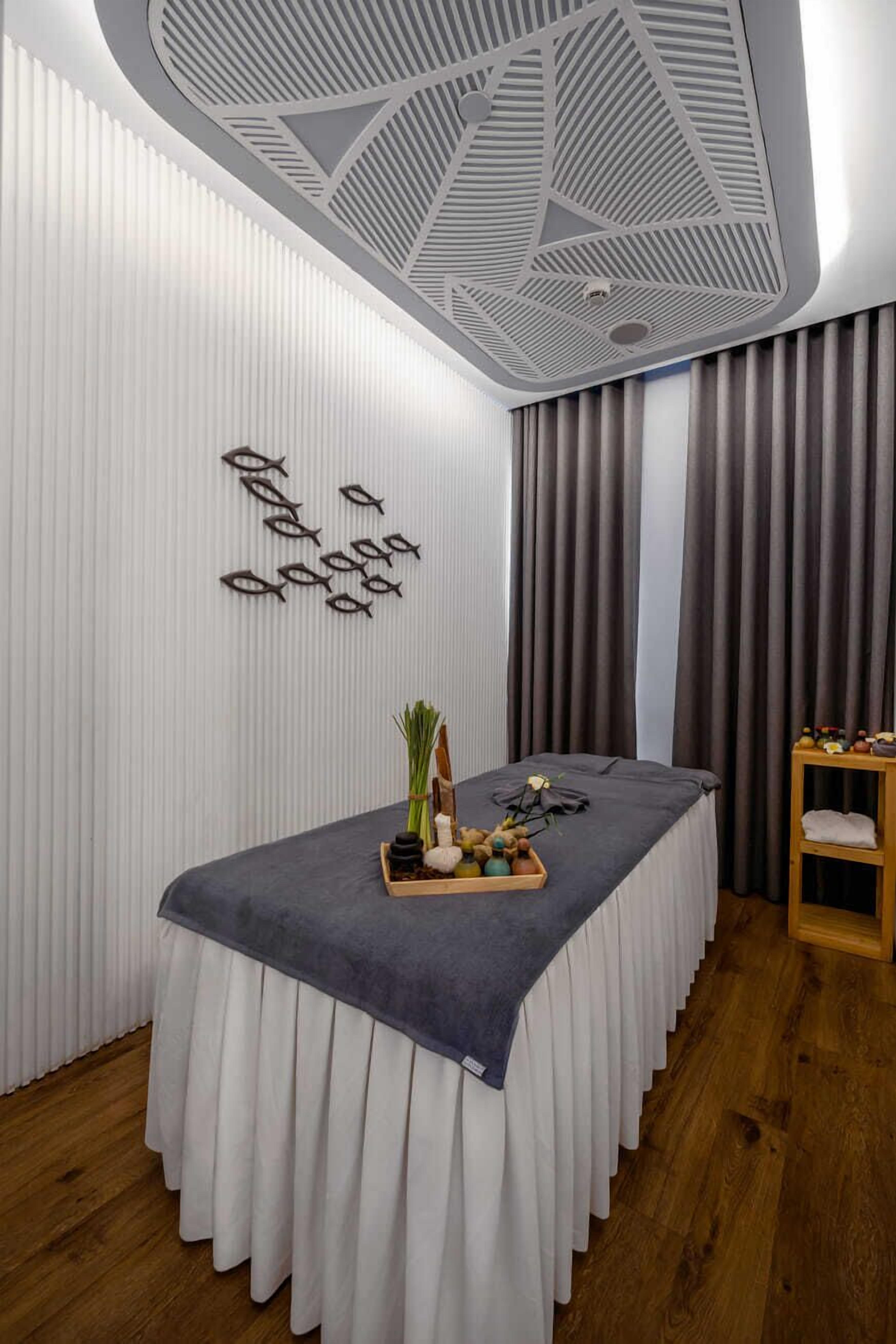 A modern spa room with a massage table, a wooden side table with towels, and a ceiling with geometric patterns.