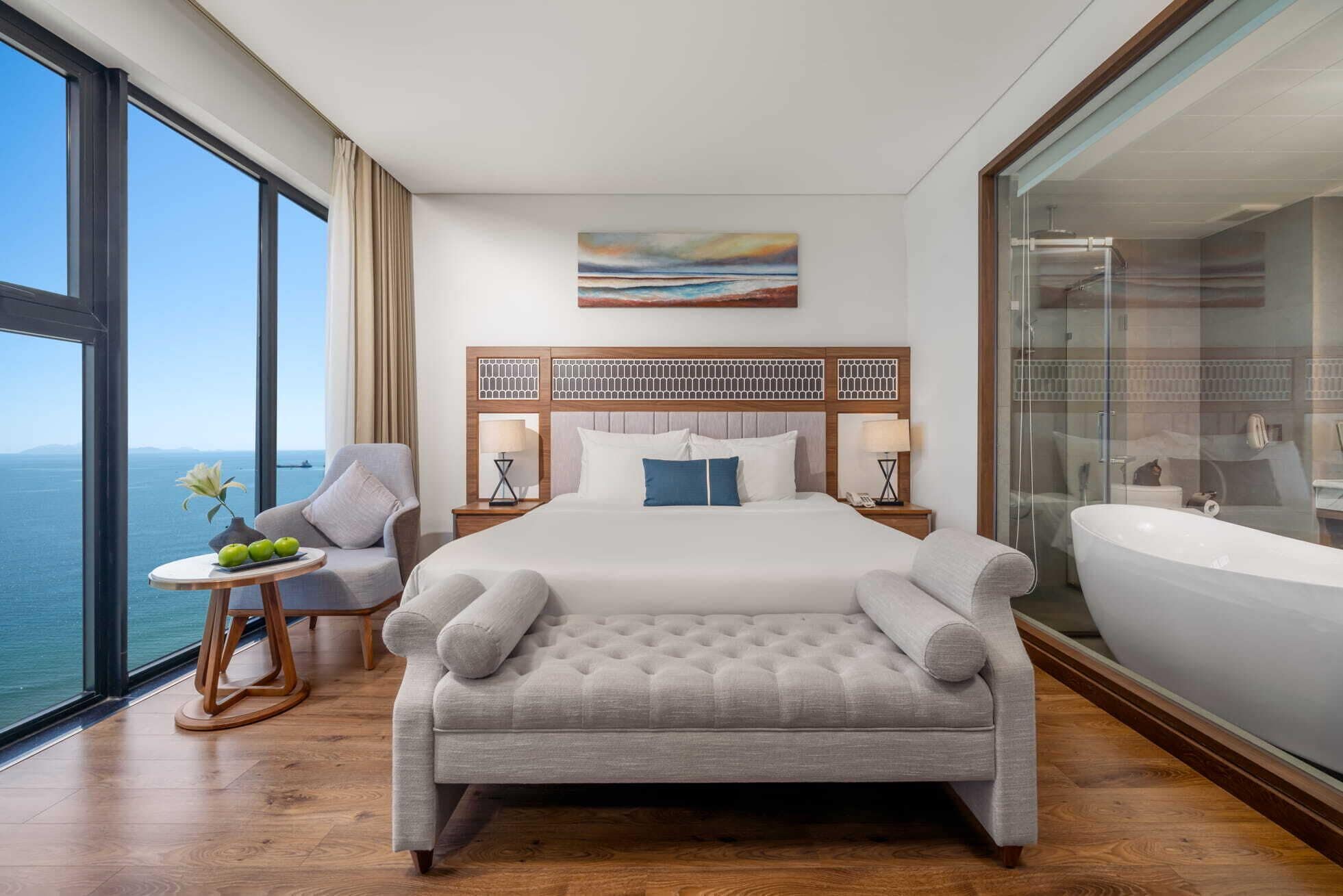 A hotel room with a large bed, a sitting area with a view of the sea, a painting on the wall, and a large bathtub.