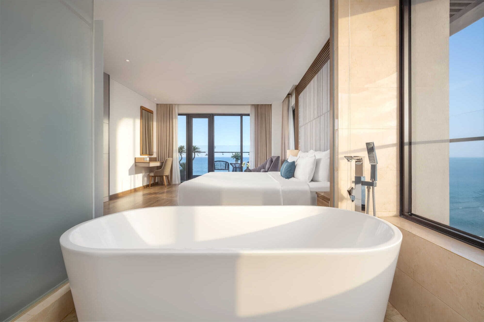 A modern hotel room with a large bathtub, a balcony with a view of the sea, and a seating area with a table and chairs.