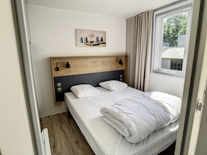 A bedroom with a wooden headboard, a bedside table with a lamp, a window with curtains, and a painting on the wall.