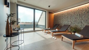 Un área de descanso moderna con dos sillones, una pequeña mesa con un florero y vistas a la ciudad a través de grandes ventanales.
