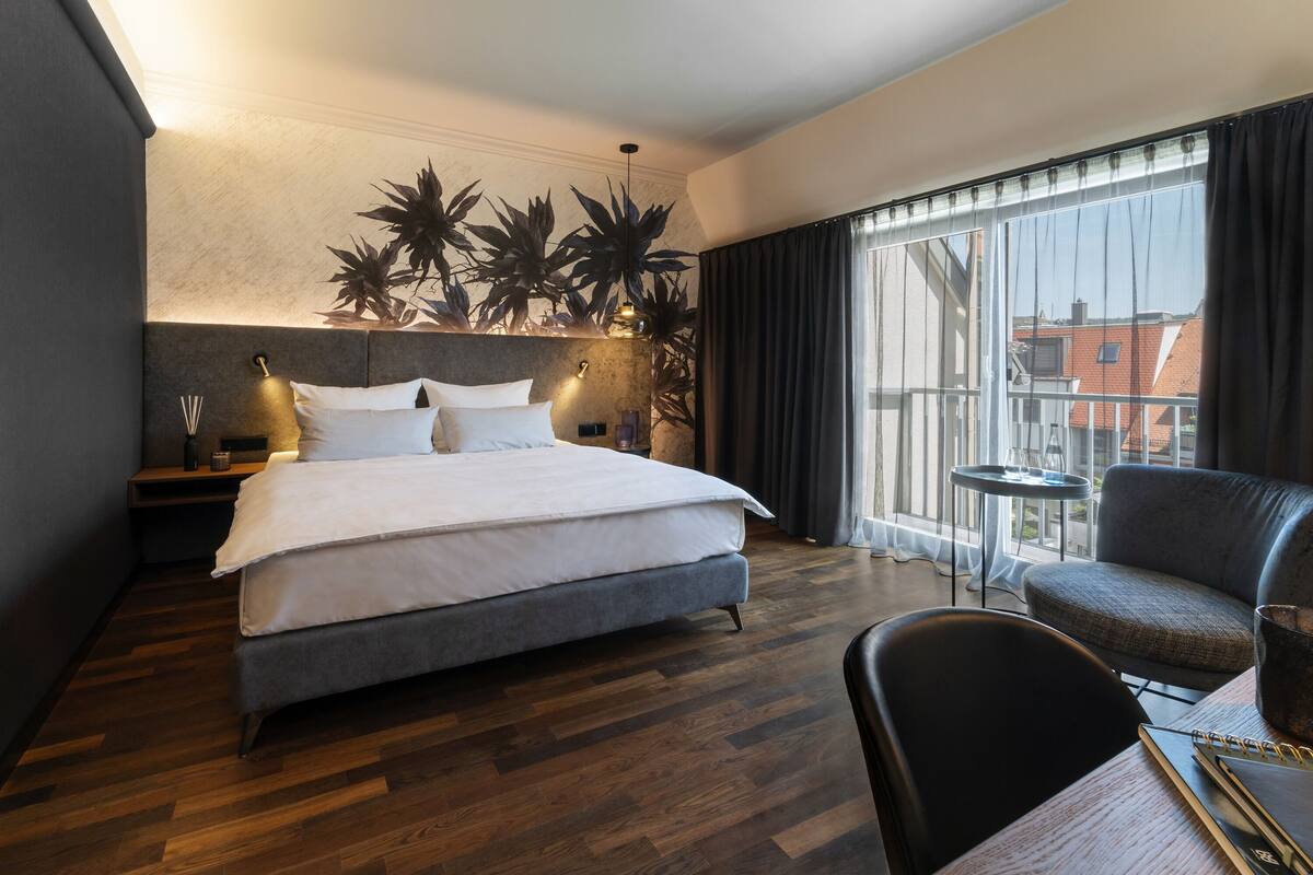 A modern hotel room with a large bed, a small table with a chair, and a balcony with a view of buildings.