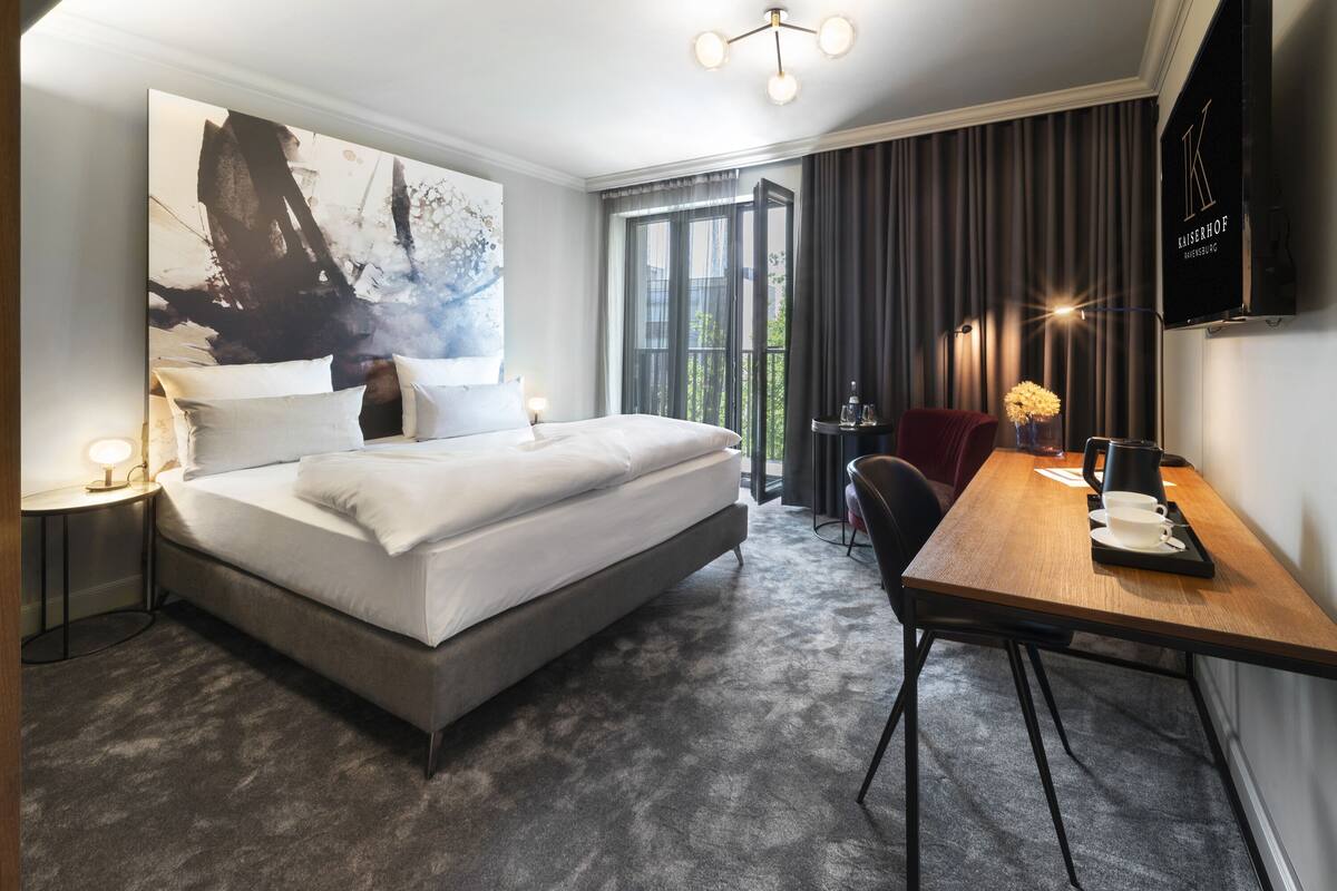 A modern hotel room with a large bed, a desk with a chair, a coffee maker, and a view of the outdoors.