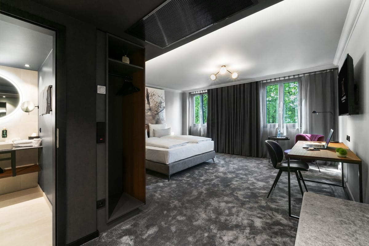 A modern hotel room with a bed, desk, and chair, a large window with curtains, and a wall-mounted TV.