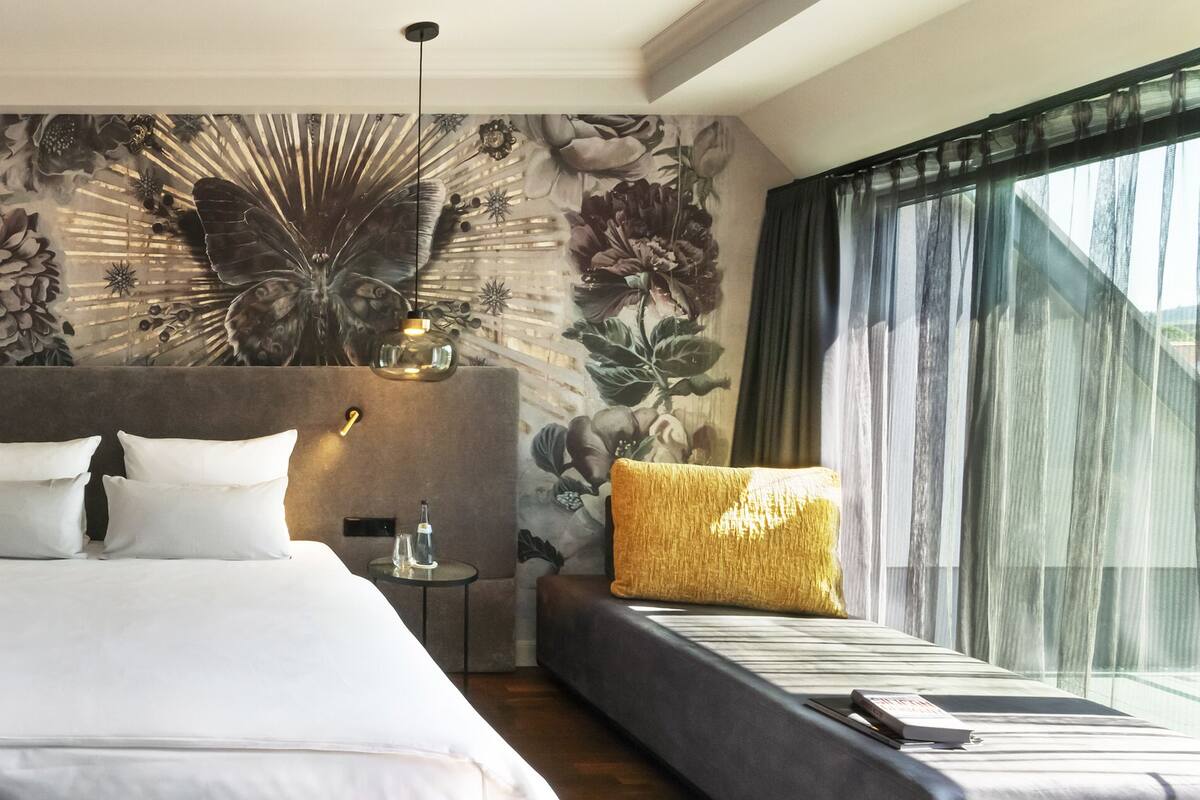 A modern hotel room with a large bed, a bedside table, a nightstand, a decorative wall mural, a large window with sheer curtains, and a view of the outdoors.
