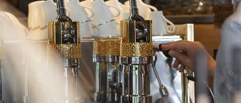 A barista preparing coffee at an espresso machine with a steam wand and portafilter, with cups and glasses in the background.