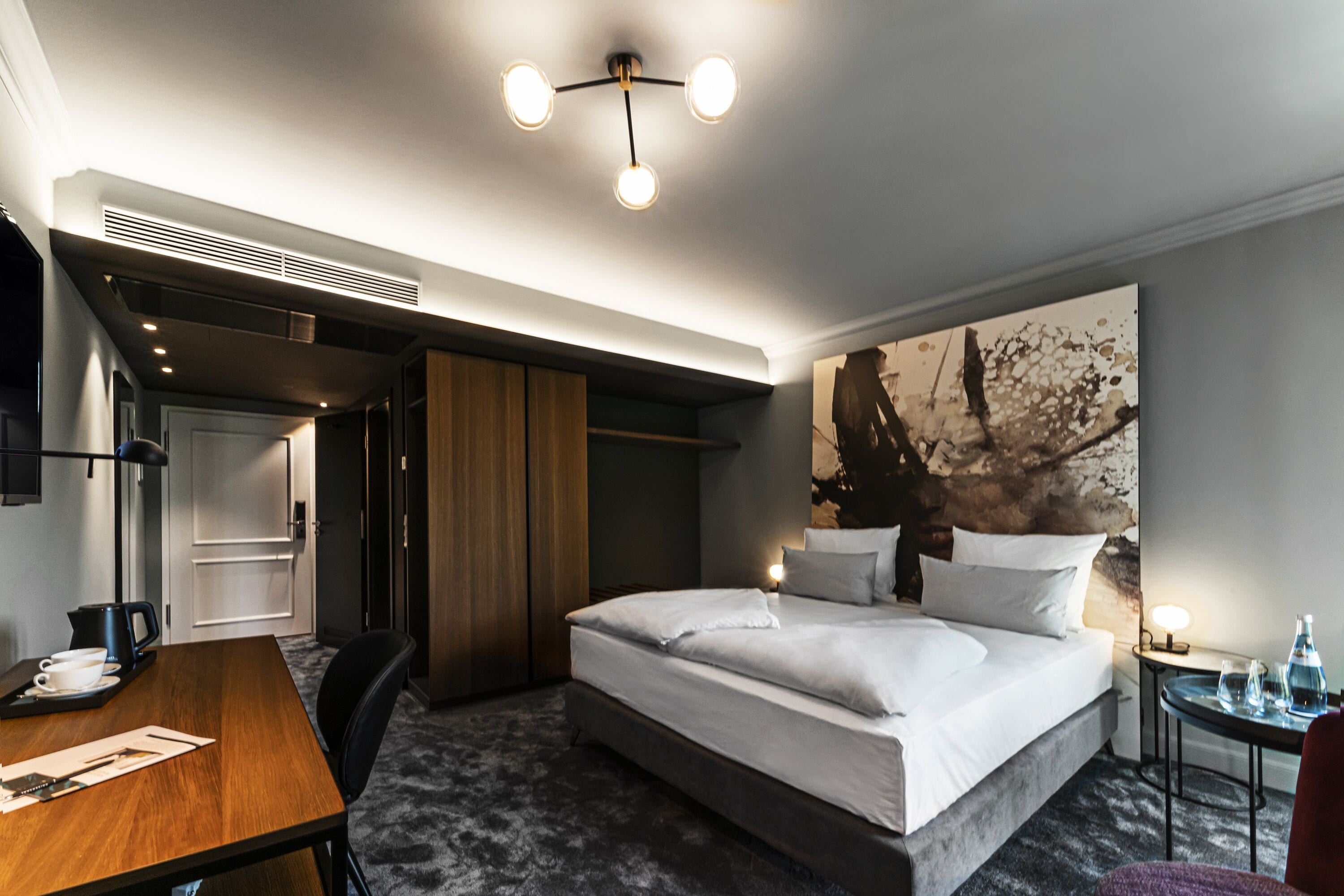 A modern hotel room with a large bed, a desk with a chair, a wardrobe, and a painting on the wall.