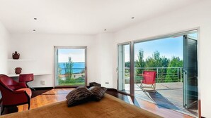 Un dormitorio moderno con una cama grande, una silla roja, una puerta de cristal que da a un balcón con otra silla roja afuera y vistas al mar.