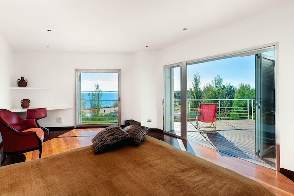 A modern bedroom with a large bed, a red chair, a glass door leading to a balcony with a red chair outside, and a view of the sea. - LA MEVA LLUNA, Luxury villa on the beachfront with pool (L'Ampolla)