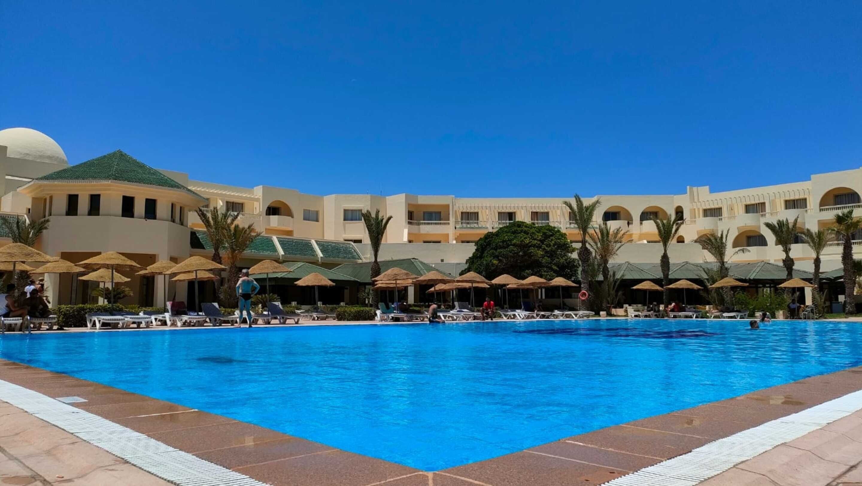 A large swimming pool with lounge chairs and umbrellas, surrounded by a hotel with multiple floors and palm trees.