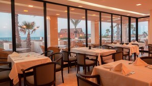 Un restaurante con mesas dispuestas para comer, amplias ventanas que ofrecen vista al mar y palmeras, y un edificio de ladrillo al fondo.