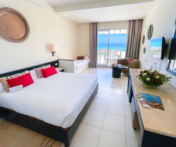 A hotel room with a large bed, a desk, a chair, a TV, and a balcony with a view of the ocean. - Vincci Dar Midoun (Djerba Midun)