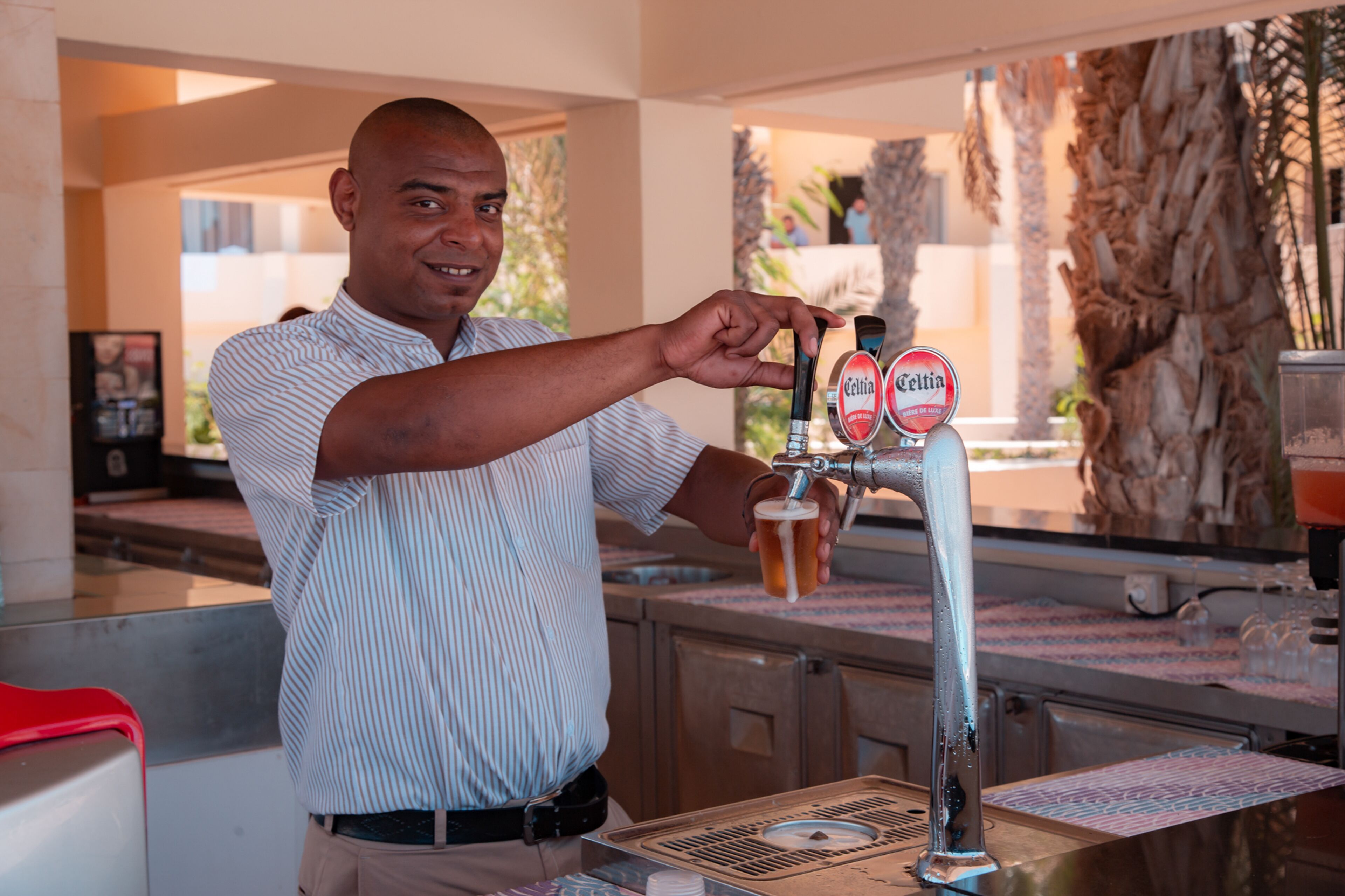 Un hombre sirviendo cerveza de una grifo con la etiqueta "Celtia" en un bar, con una barra y una palmera al fondo.