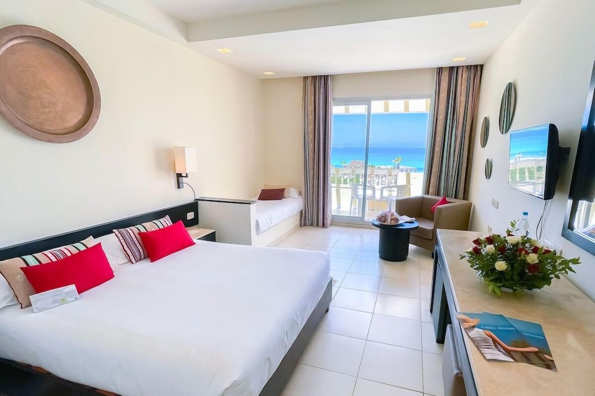 A hotel room with a bed, a desk, a chair, a TV, and a view of the ocean.