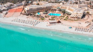 Vista aérea de un hotel costero con una amplia piscina, zona de playa con tumbonas y varios edificios con balcones.