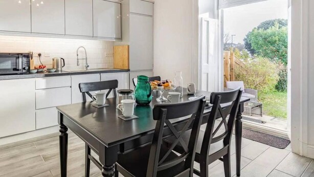 A modern kitchen with white cabinets, a dining table with chairs, and a view of the garden through a large window. - Lovely Villa 1 Double 1 Double Sofa 6 Single,1bunk (Torquay)