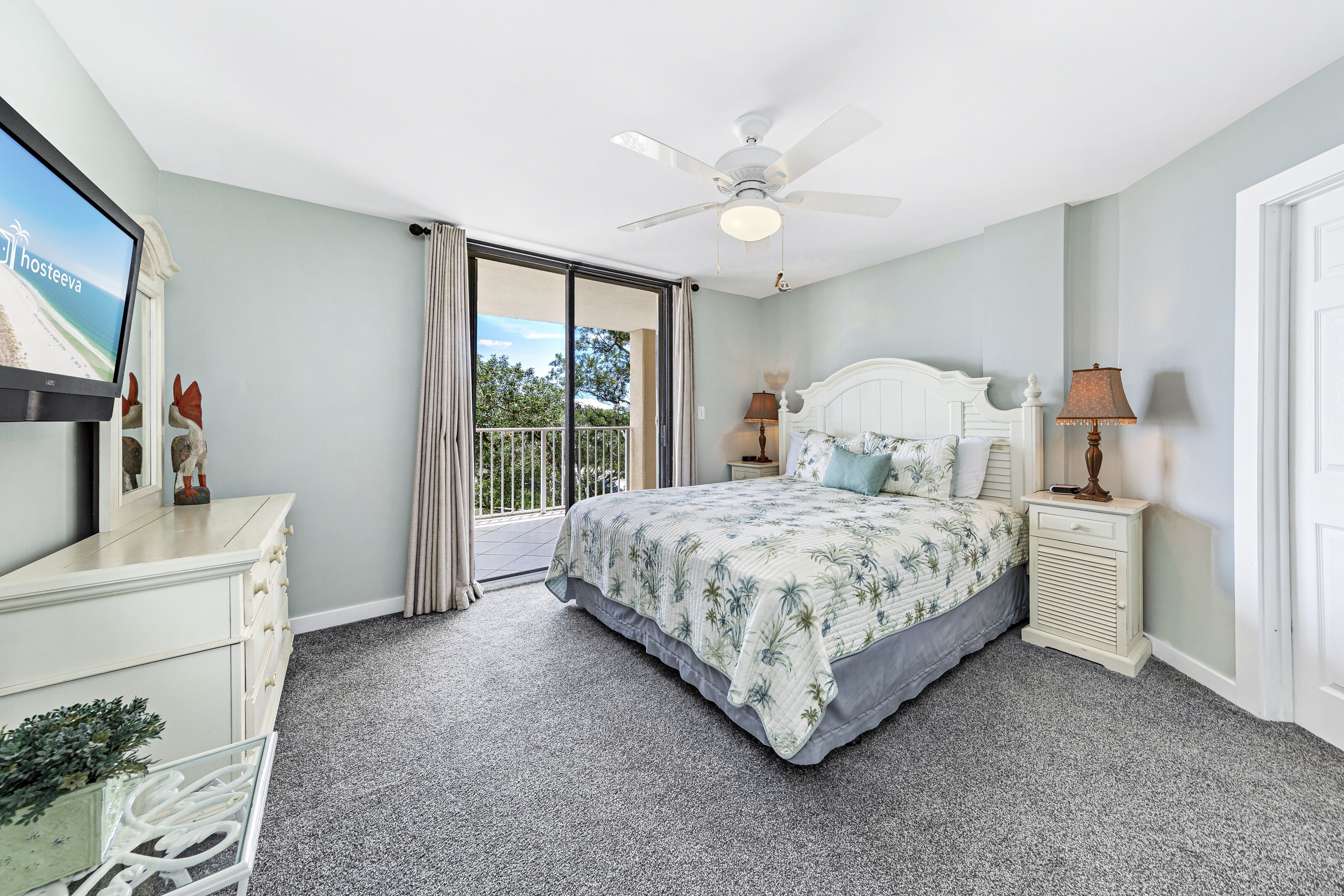 Une chambre à coucher comprenant un lit, une table de chevet, une lampe, un ventilateur de plafond, un téléviseur et une porte-fenêtre coulissante donnant sur un balcon.