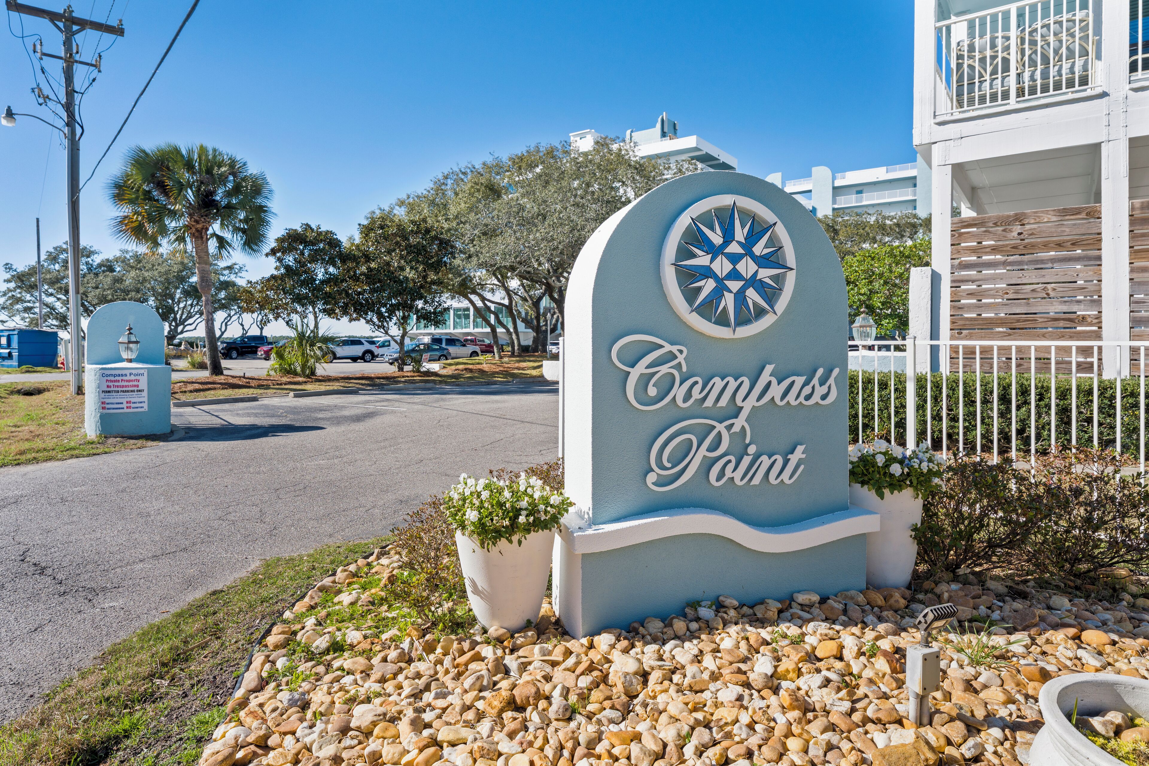 Letrero de Compass Point con un emblema decorativo de estrella, rodeado de piedras y plantas en macetas.