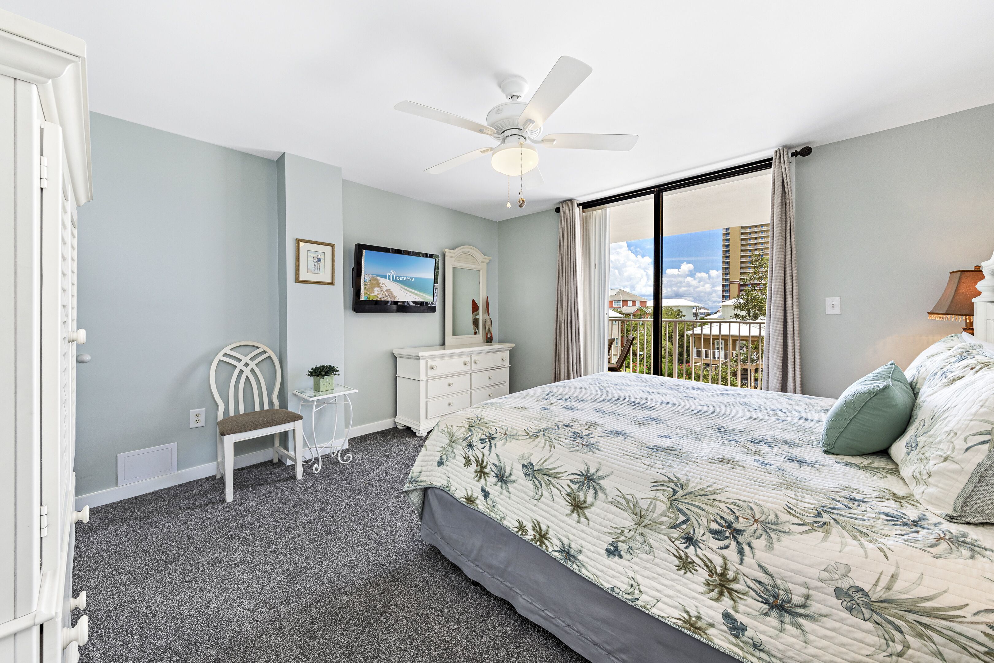 A bedroom with a bed, a chair, a dresser, a ceiling fan, and a window with a view of buildings.