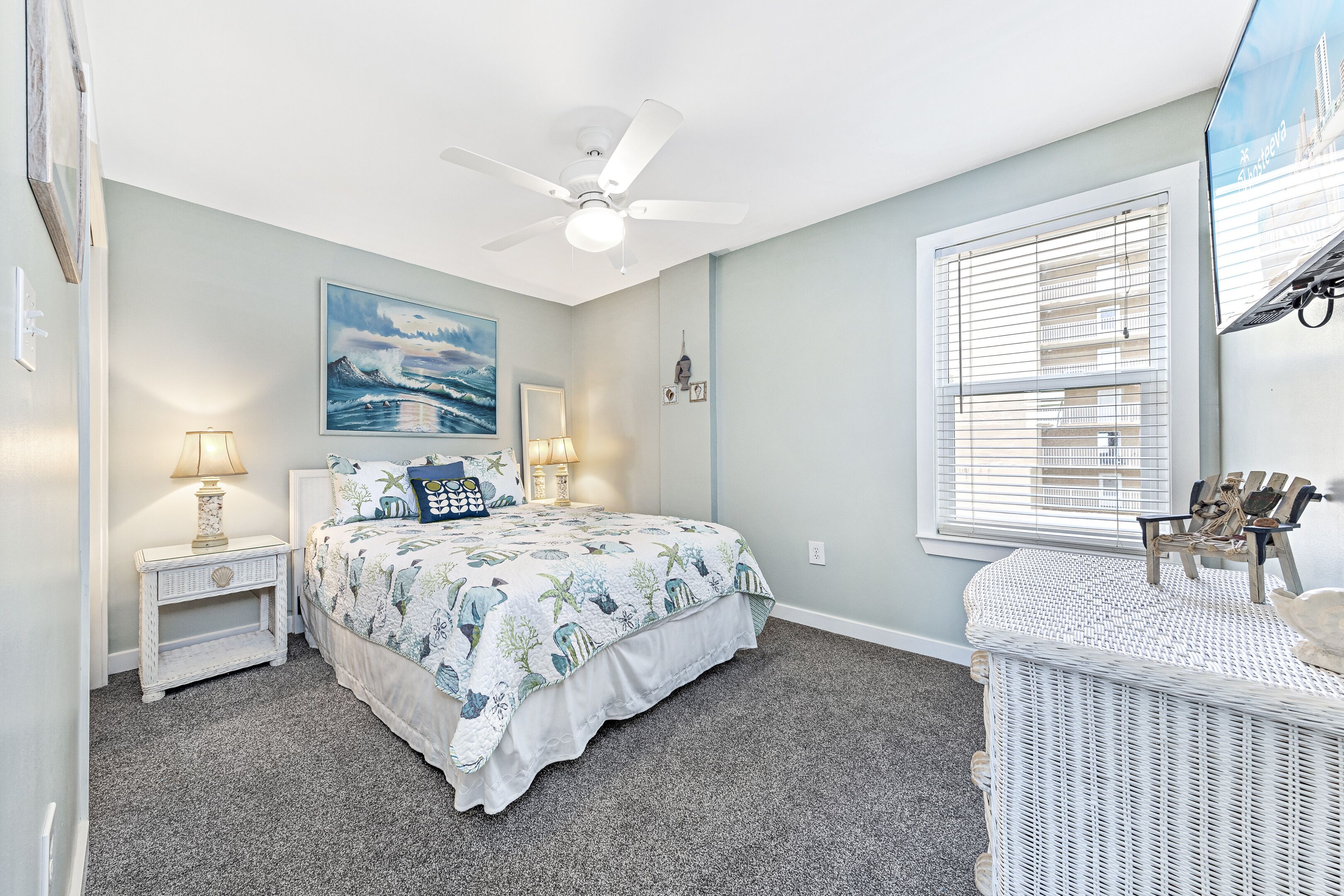 Une chambre à coucher comprenant un lit, des tables de chevet, un ventilateur de plafond, une fenêtre avec des stores et un fauteuil en osier.
