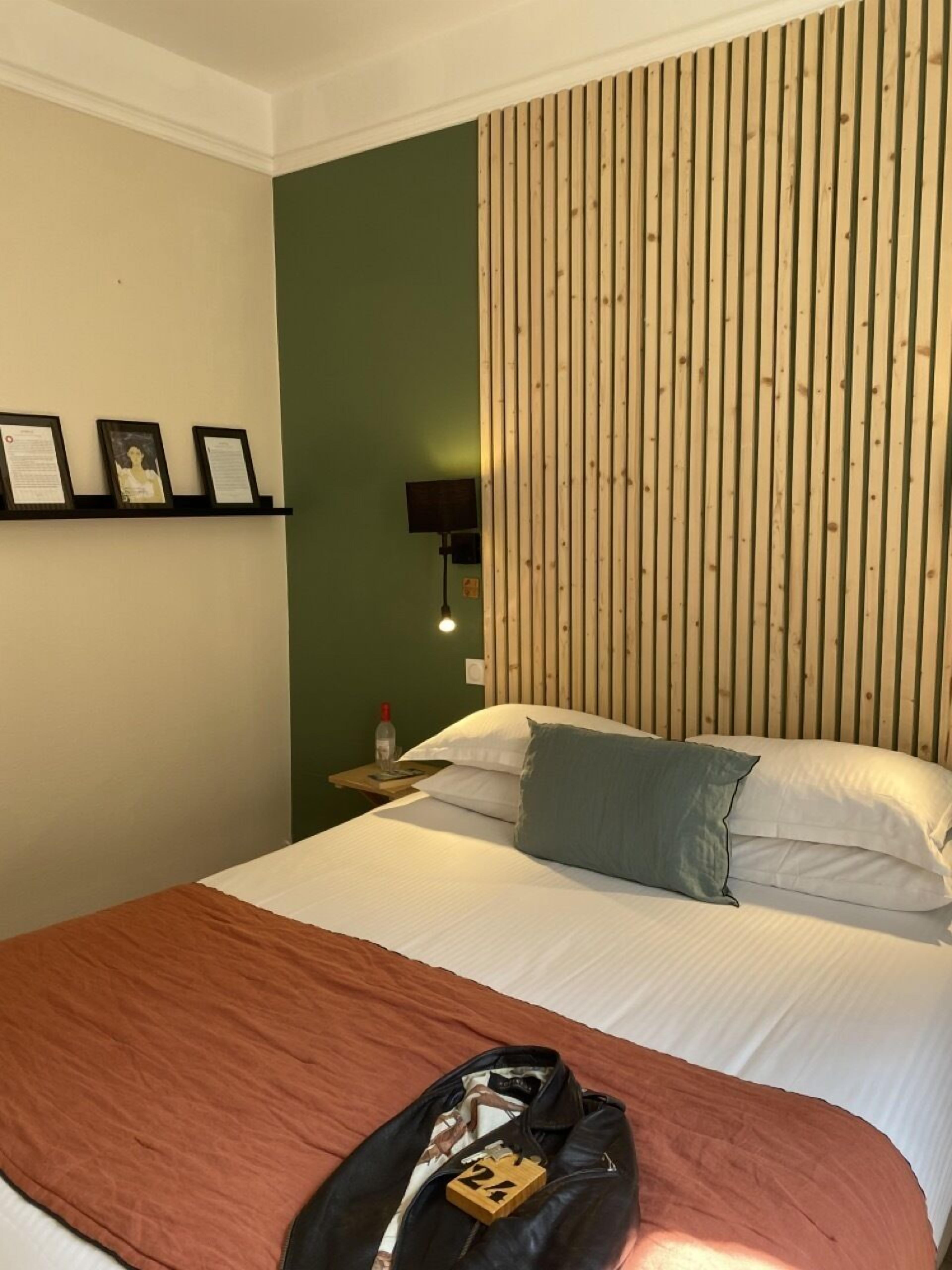 A hotel room with a bed, a nightstand, a wall-mounted shelf with framed items, and a wooden slat wall.