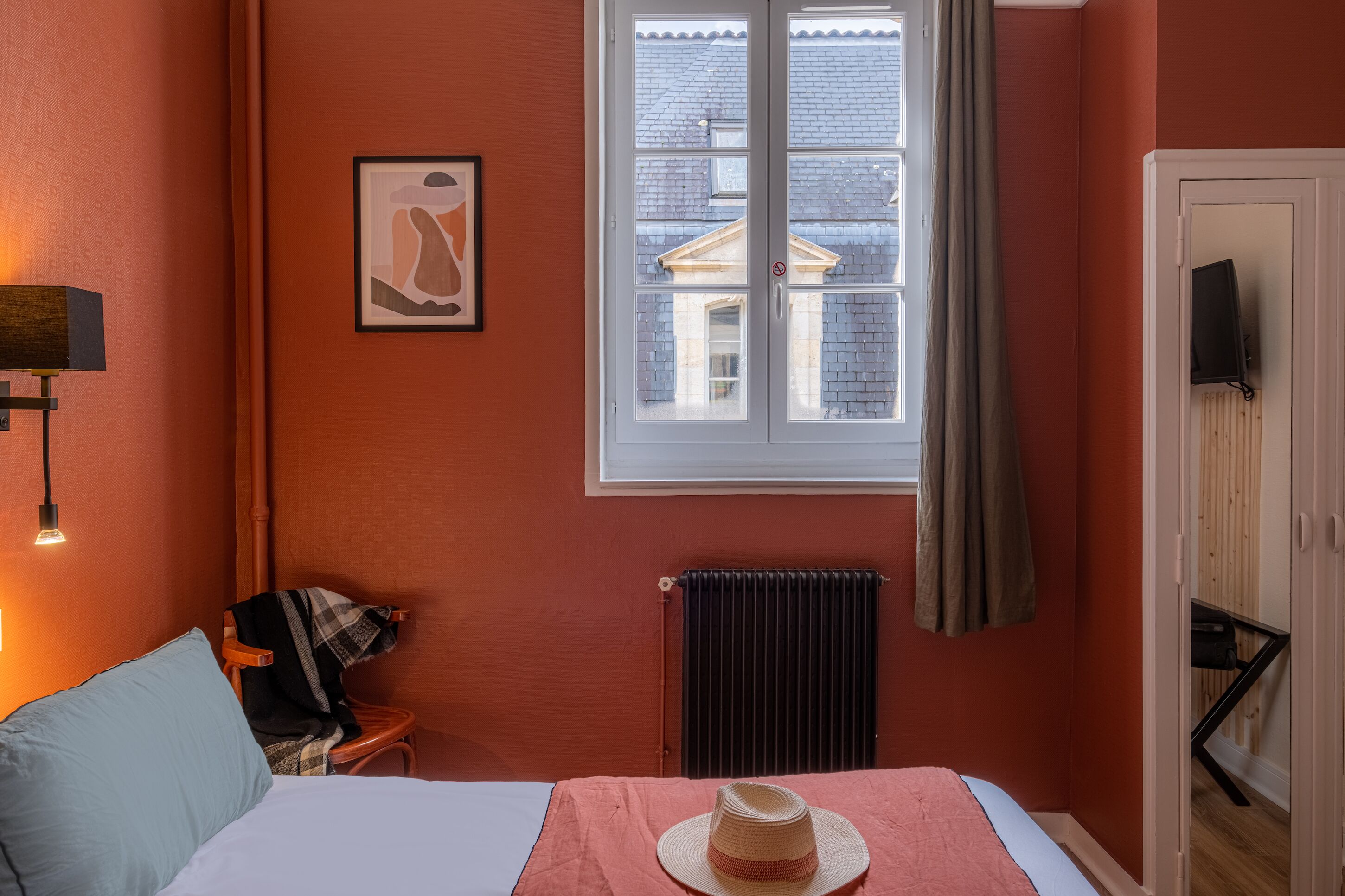 Una habitación con una cama, un radiador, un ventanal con cortinas, un cuadro en la pared y una lámpara.