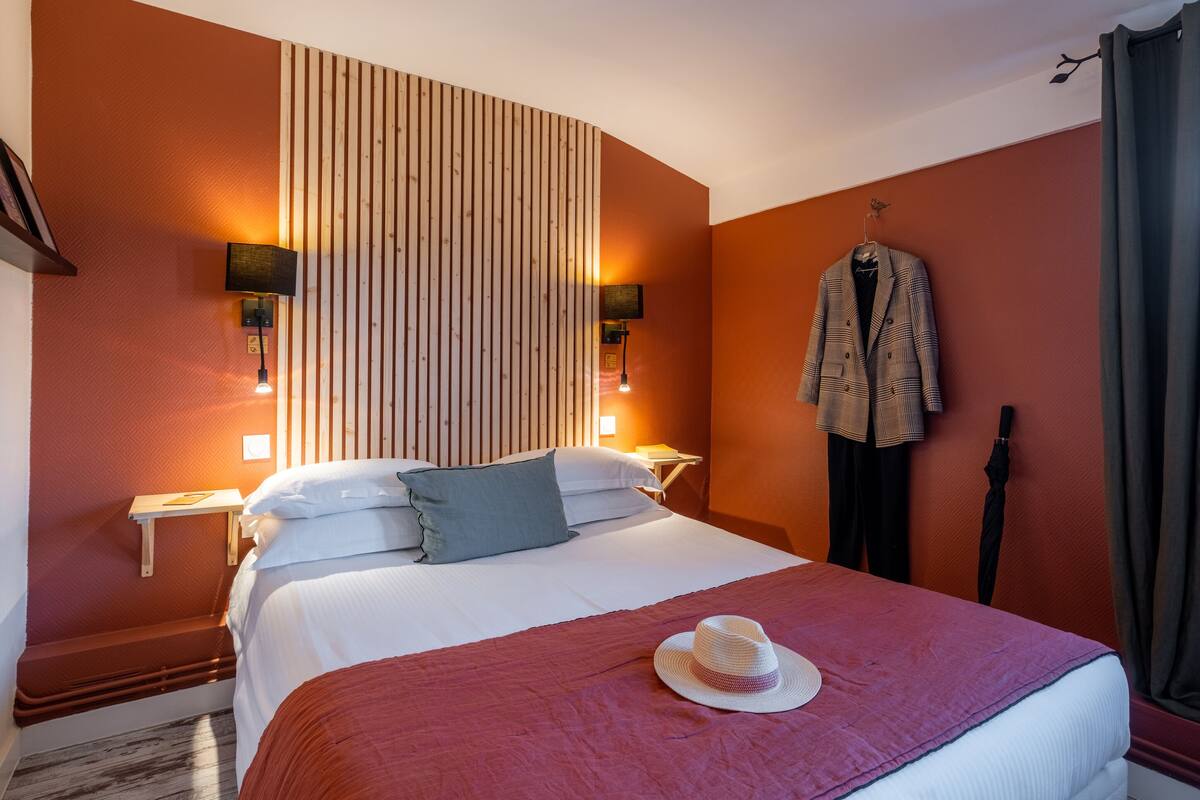 A neatly made bed with a red blanket, a white pillow, and a hat on the bed. There are two wall-mounted lamps on either side of the bed.