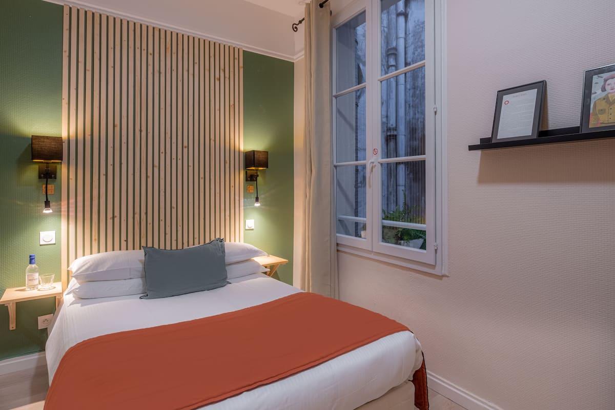 A neatly arranged bedroom with a bed, bedside tables, a window, and a shelf with framed pictures.