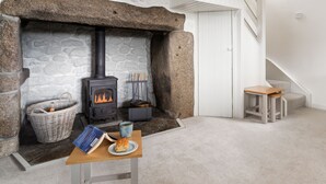 Una habitación acogedora con una chimenea de piedra, una cesta de mimbre, una mesa con un libro y un plato de comida, y un taburete de madera.