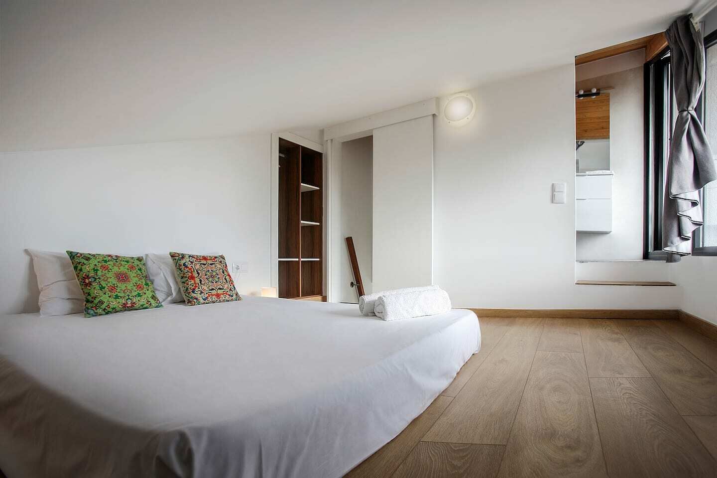 A bedroom with a large bed, a wooden wardrobe, a window with curtains, and a built-in shelf.