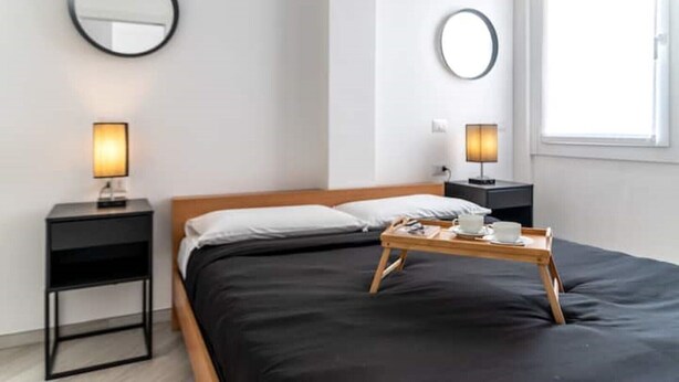 Una camera d'albergo moderna con un letto, due comodini, una lampada da comodino, un vassoio con tazza e piattino, e una finestra con luce naturale.