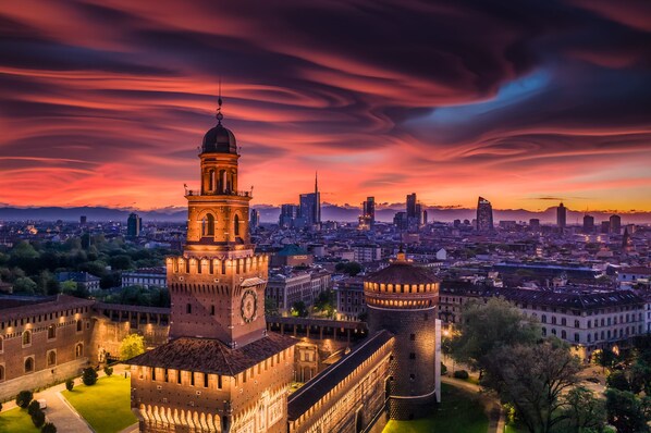 A historic building with a prominent clock tower and a round tower, surrounded by a cityscape at sunset.