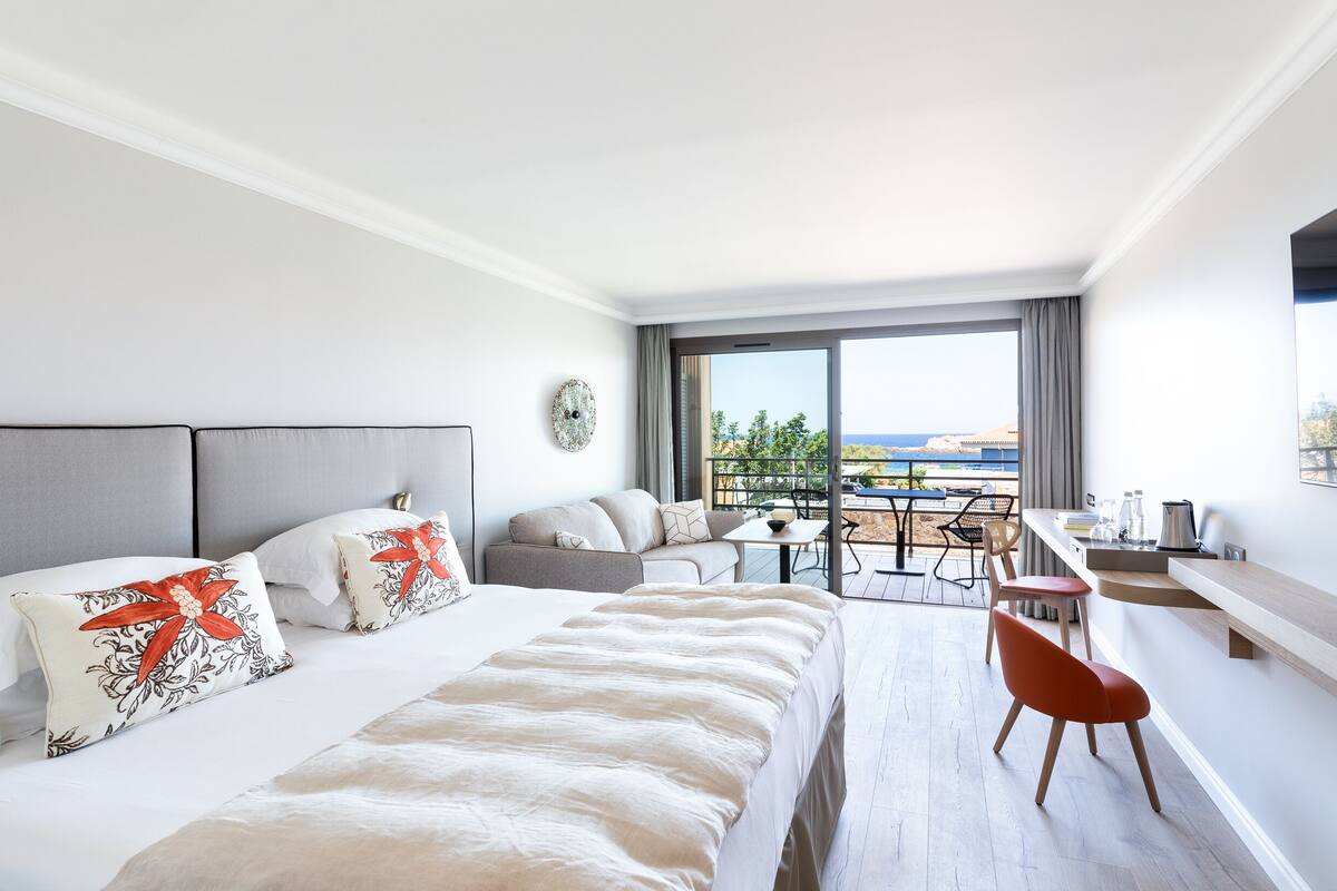 A modern hotel room with a large bed, a sofa, a dining table, and a balcony with a view.