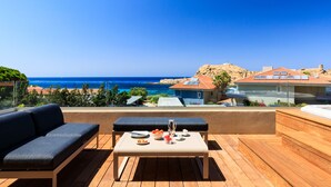 A wooden deck with a sofa and table, overlooking a coastal view with buildings and a clear sky.