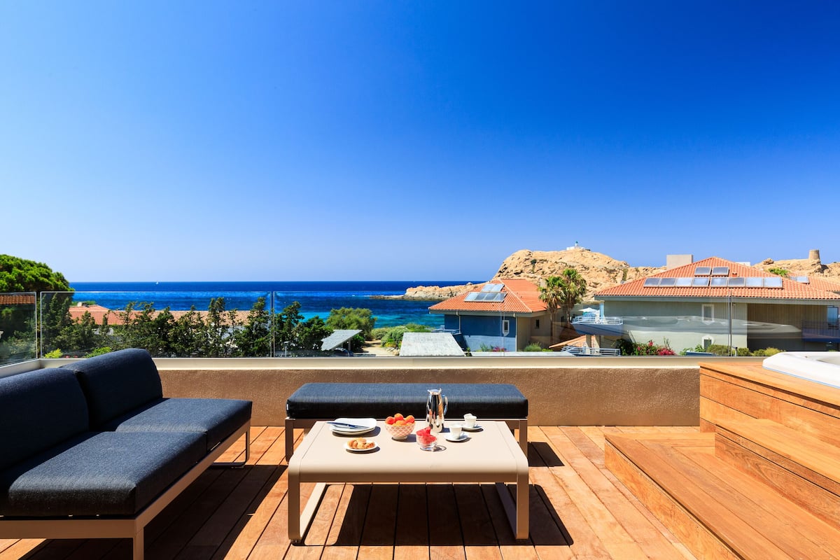 A wooden deck with a sofa and table, overlooking a coastal view with buildings and a clear sky.