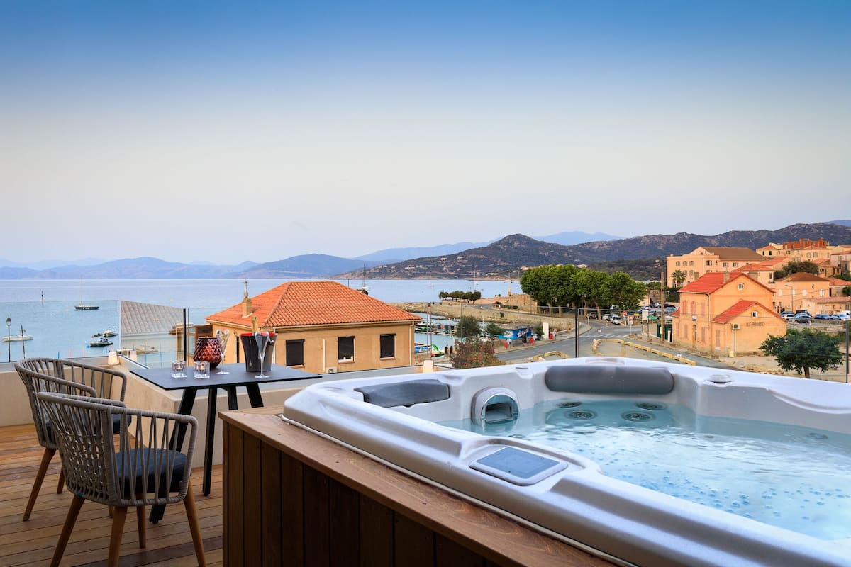 A hot tub on a wooden deck with a table and chairs, overlooking a coastal town with buildings and boats.